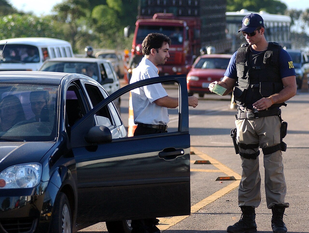 Agente da Policia Rodoviaria Federal em blitz de fiscalizacao em rodovia federal, com caminhao de carga ao fundo